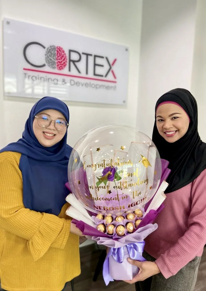 Amal Azimah Zahidah Ali Ajis and Azrinna Kepli holding a congratulatory bouquet in front of the Cortex Training and Development sign