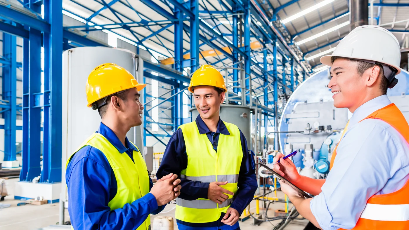 Three workers in hard hats and high-visibility vests talking together in a plant, one holding a clipboard.