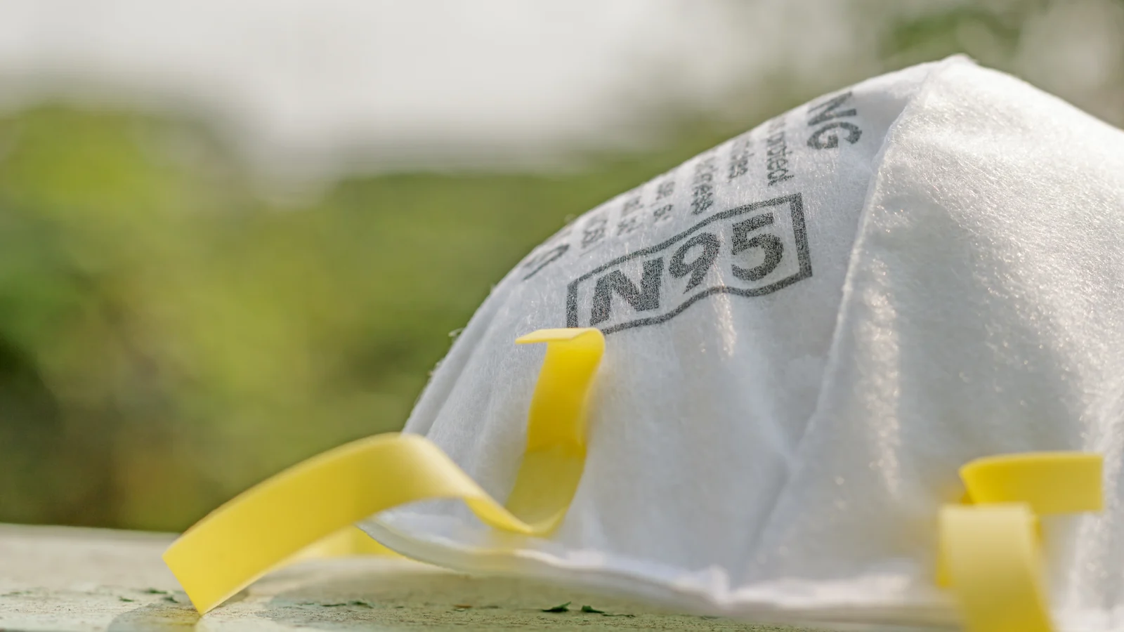 A white N95 respirator with yellow head straps, resting outdoors against blurred greenery
