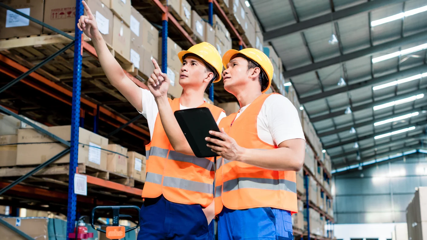 Two warehouse workers in high-visibility vests and hard hats looking up at high storage racking, one holding a tablet.