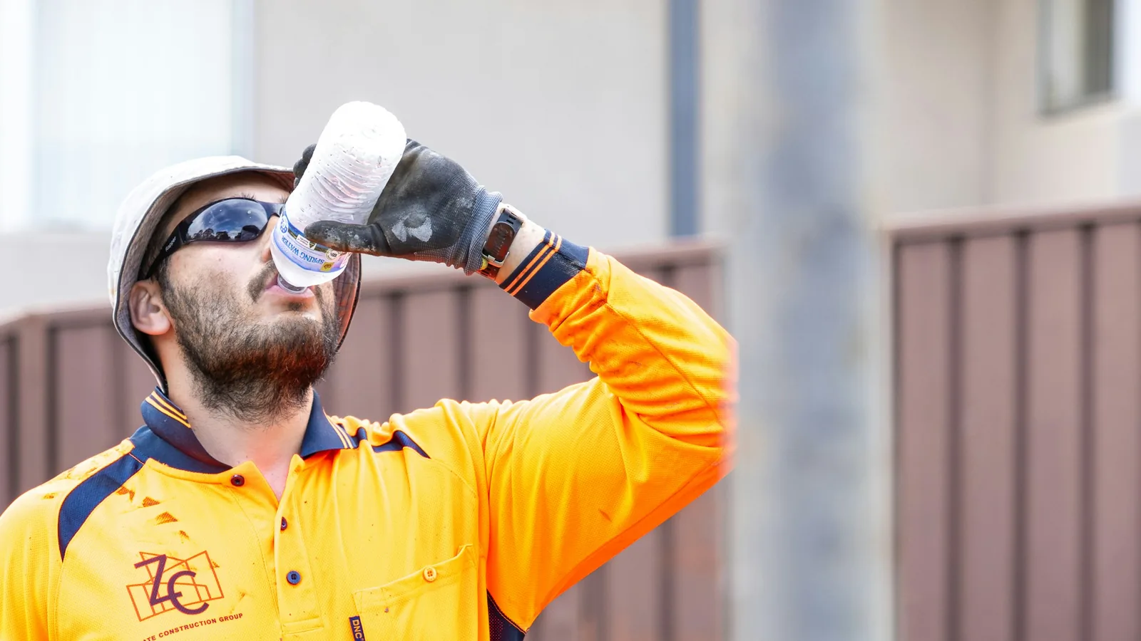 A construction worker in an orange high-visibility shirt, hard hat and gloves drinking from a water bottle on site